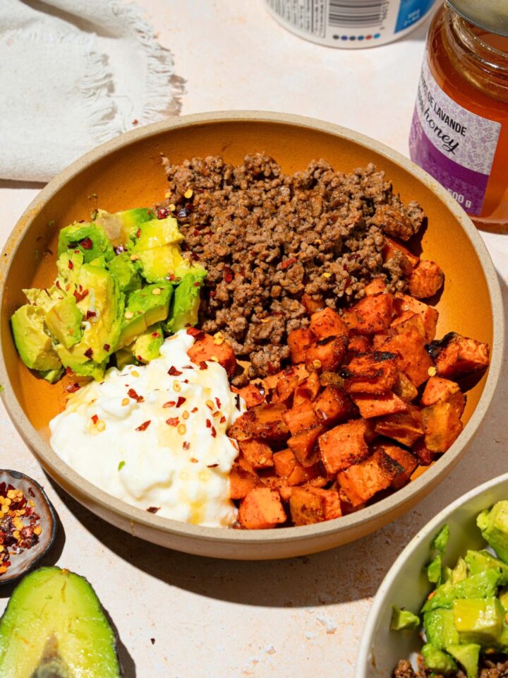 A bowl that is filled with ground beef, diced, roasted sweet potatoes, cottage cheese, and sliced avocado with hot honey drizzled on top.