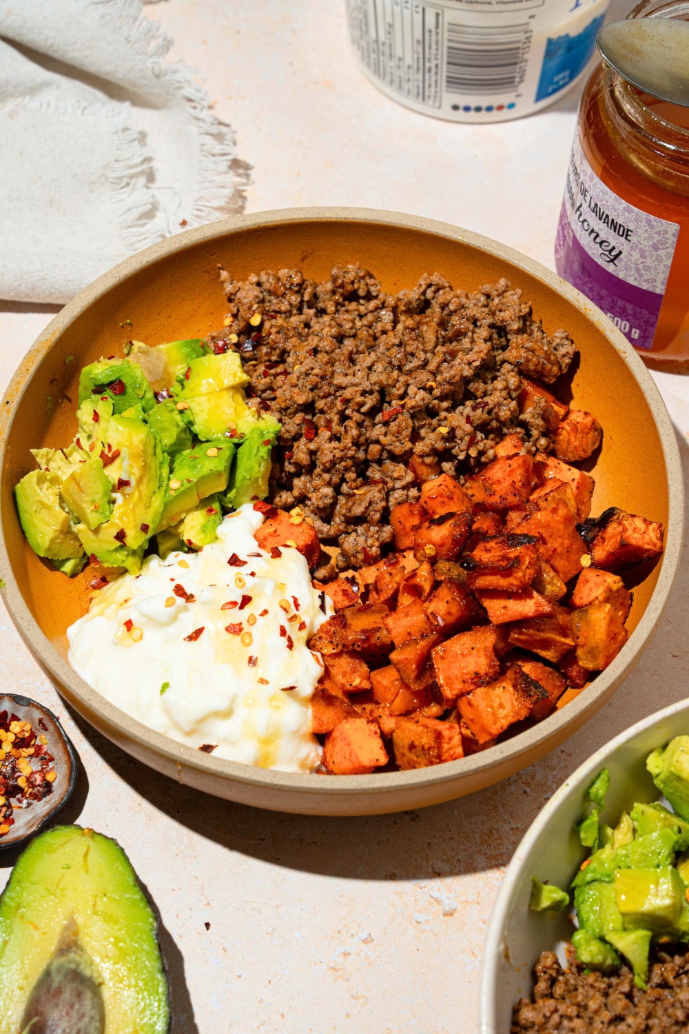 A bowl that is filled with ground beef, diced, roasted sweet potatoes, cottage cheese, and sliced avocado with hot honey drizzled on top.