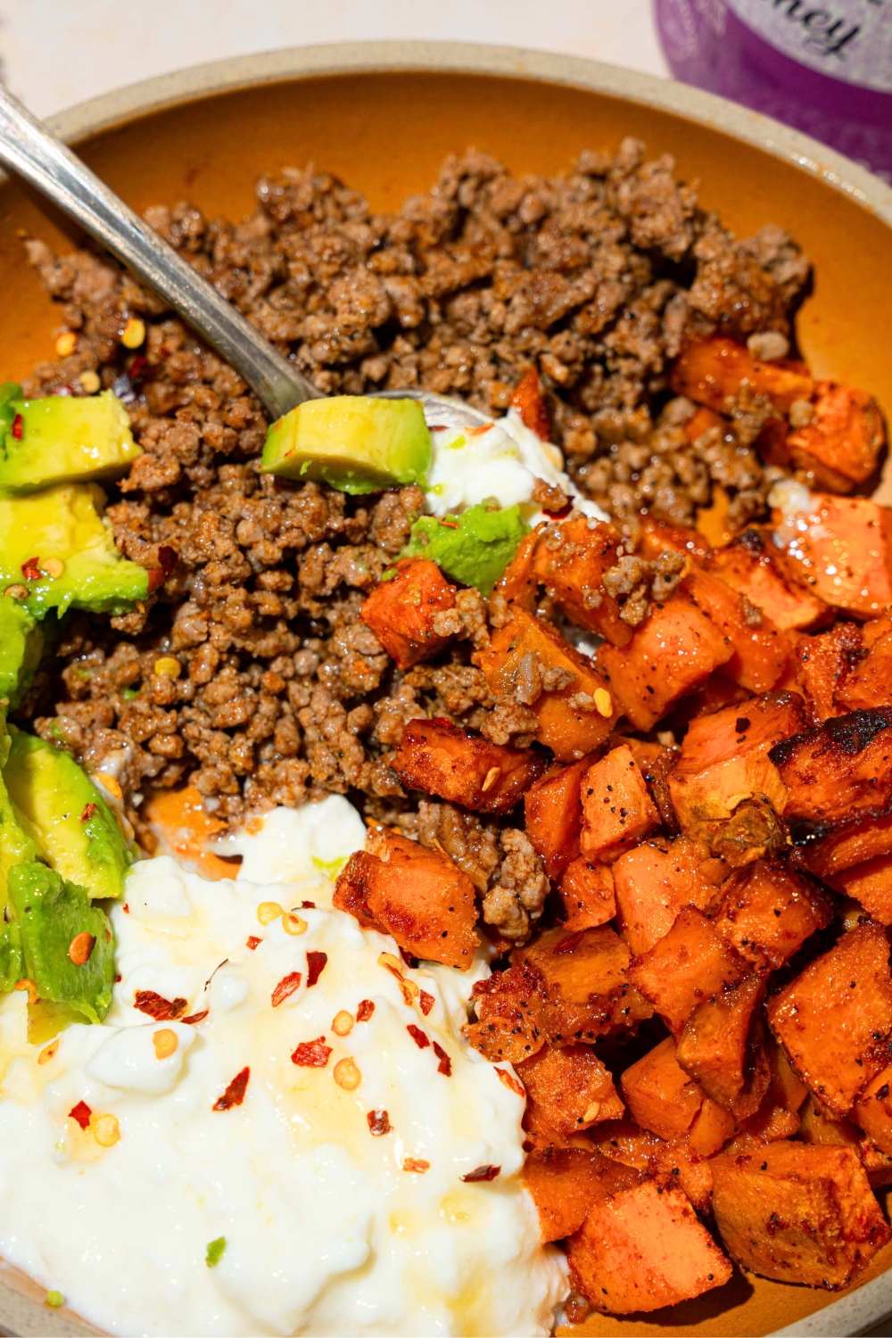 A sweet potato ground beef bowl that has sweet potatoes and ground beef, cottage cheese, avocado, hot, honey, and red pepper flakes.