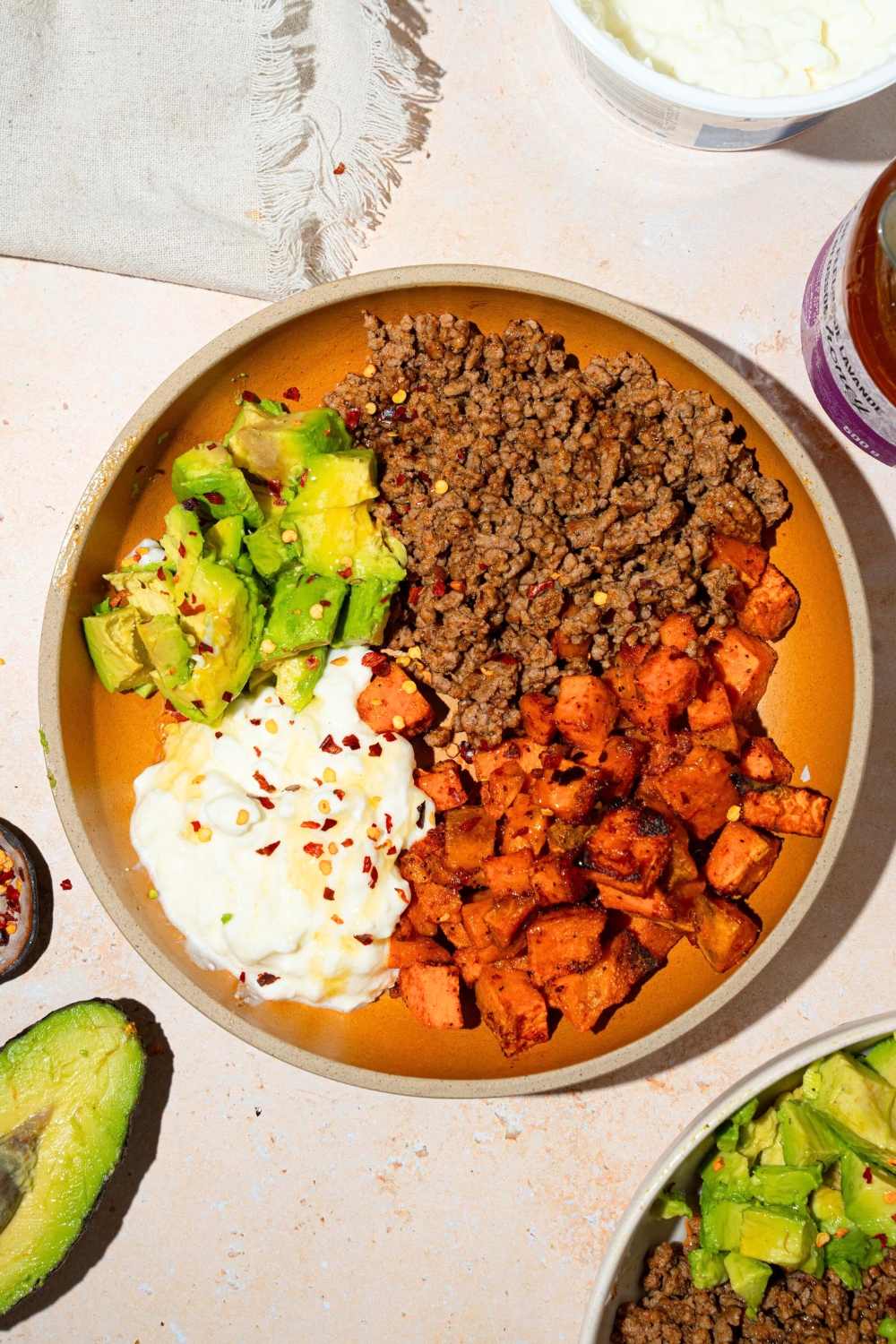 Hot honey and red pepper flakes on top of dice, roasted sweet potatoes, brown, ground beef, diced avocado, and cottage cheese, all separated in the sections in a bowl.