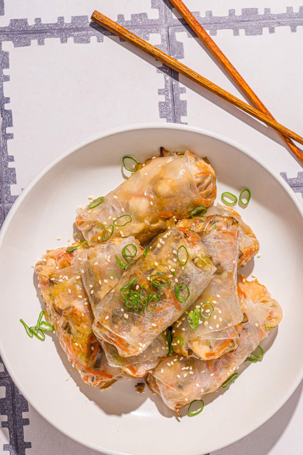 A white bowl with vegetable rice paper dumplings garnished with sliced green onions and sesame seeds. The dumplings are lightly fried. The bowl is on a tiled counter with a pair of chopsticks and small.