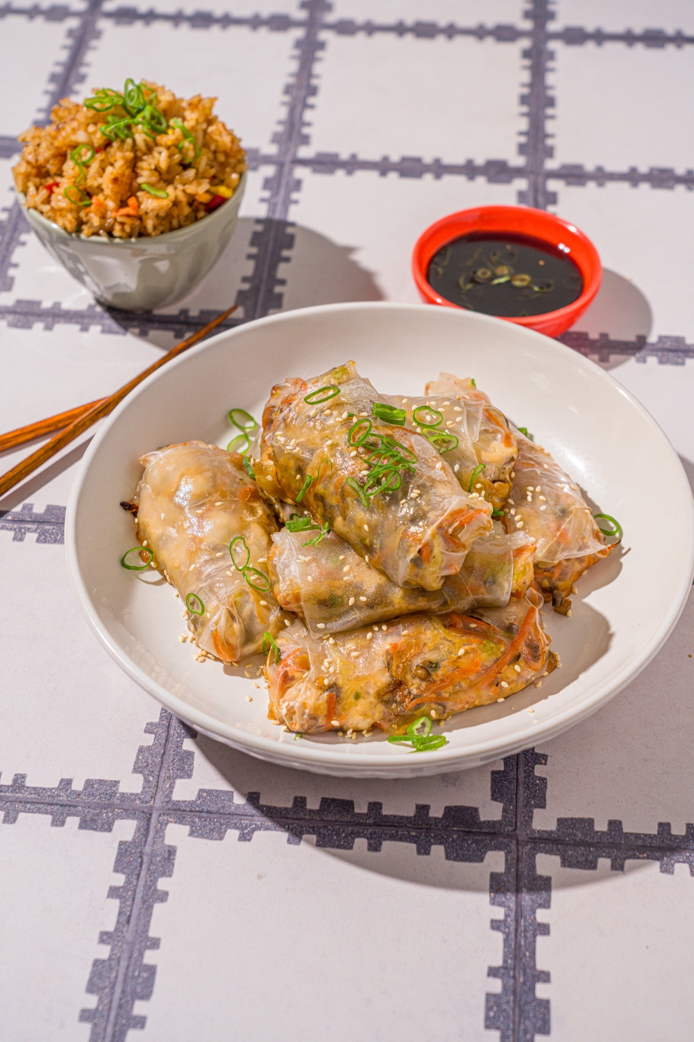 A white bowl with vegetable rice paper dumplings garnished with sliced green onions and sesame seeds. The dumplings are lightly fried. The bowl is on a tiled counter with a pair of chopsticks and small bowl of dipping sauce.