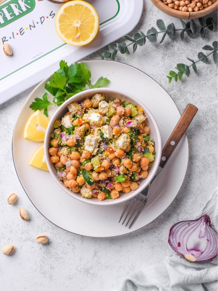 The Jennifer Aniston salad consisting of quinoa, chickpeas, cucumber, onion, feta cheese, pistachios, and mint tossed in a lemon olive oil dressing in a white bowl on top of a white plate that is on a white counter.