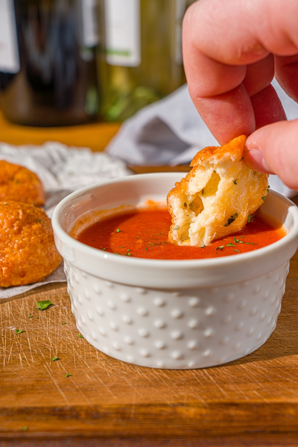 A hand dipping an air fryer parmesan cheese ball into a small ramekin of marinara sauce. The ramekin is on a wooden counter with additional cheese balls.