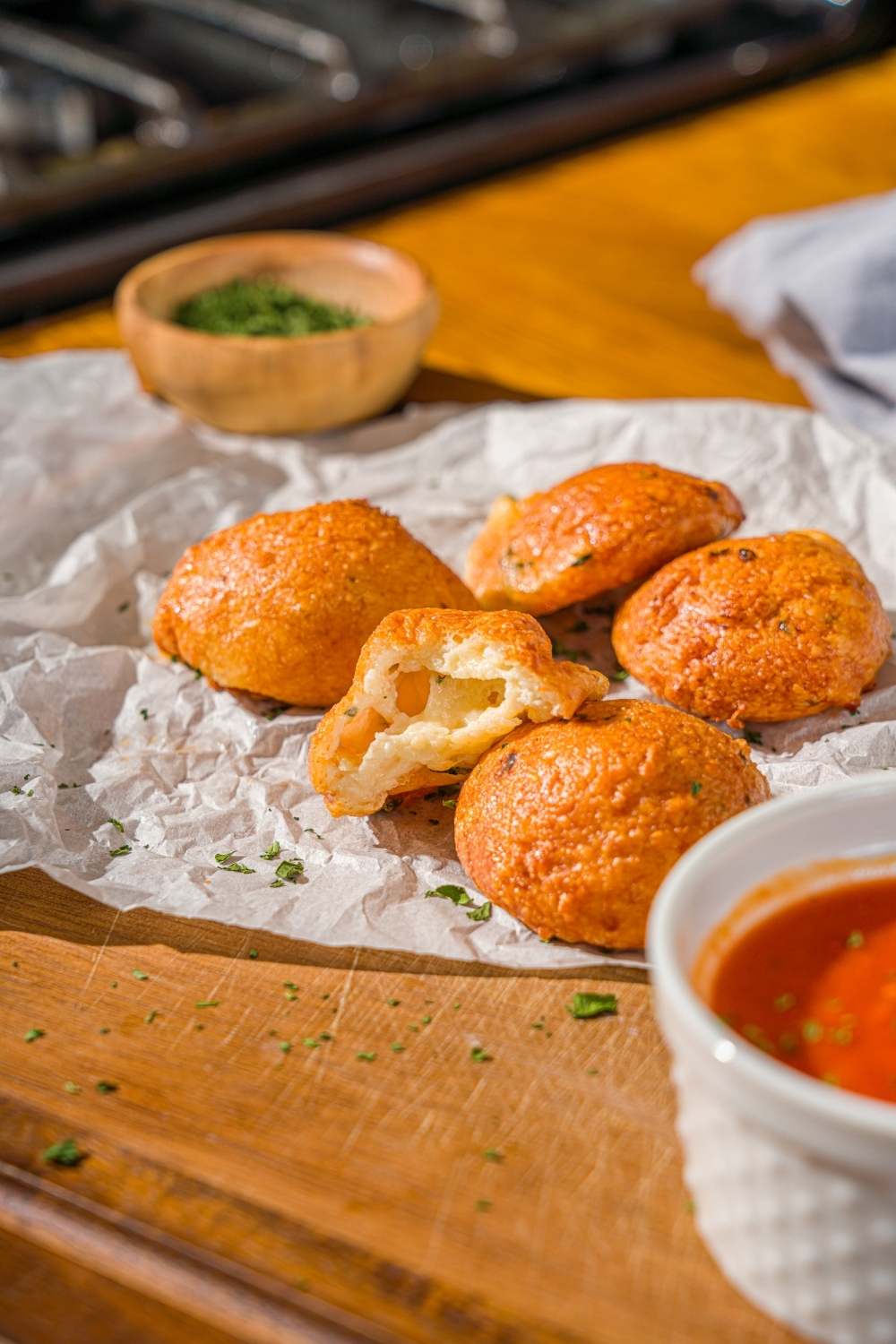 Several air fryer parmesan cheese balls on a piece of parchment paper on a wooden counter. A bite is taken from one of the cheese balls. There is a small bowl of parsley and ramekin of marinara sauce on the counter.