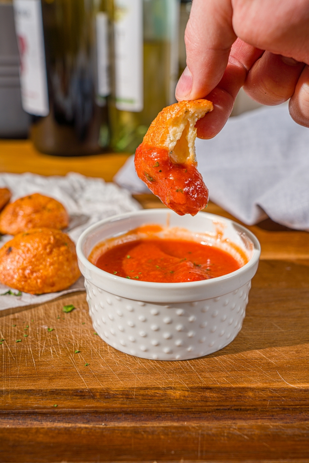 A hand dipping an air fryer parmesan cheese ball into a small ramekin of marinara sauce. The ramekin is on a wooden counter with additional cheese balls.
