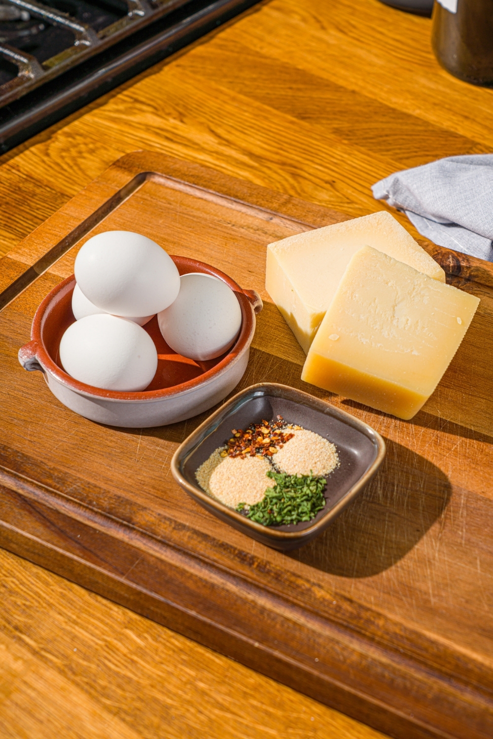 A wooden cutting board with ingredients to make air fryer parmesan cheese balls including eggs, parmesan cheese blocks, and seasonings.