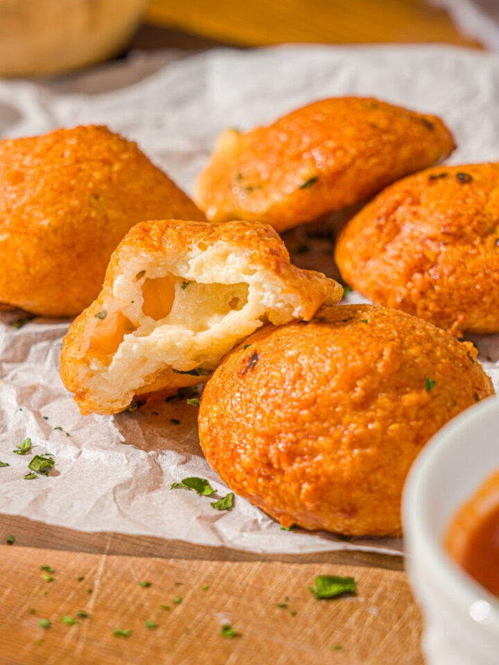 Several air fryer parmesan cheese balls on a piece of parchment paper on a wooden counter. A bite is taken from one of the cheese balls. There is a small bowl of parsley and ramekin of marinara sauce on the counter.