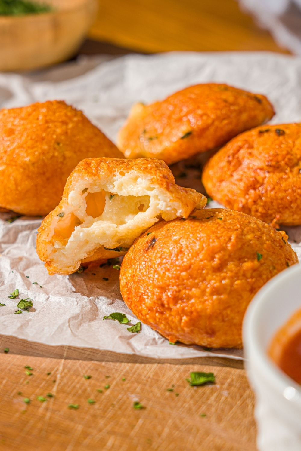 Several air fryer parmesan cheese balls on a piece of parchment paper on a wooden counter. A bite is taken from one of the cheese balls. There is a small bowl of parsley and ramekin of marinara sauce on the counter.
