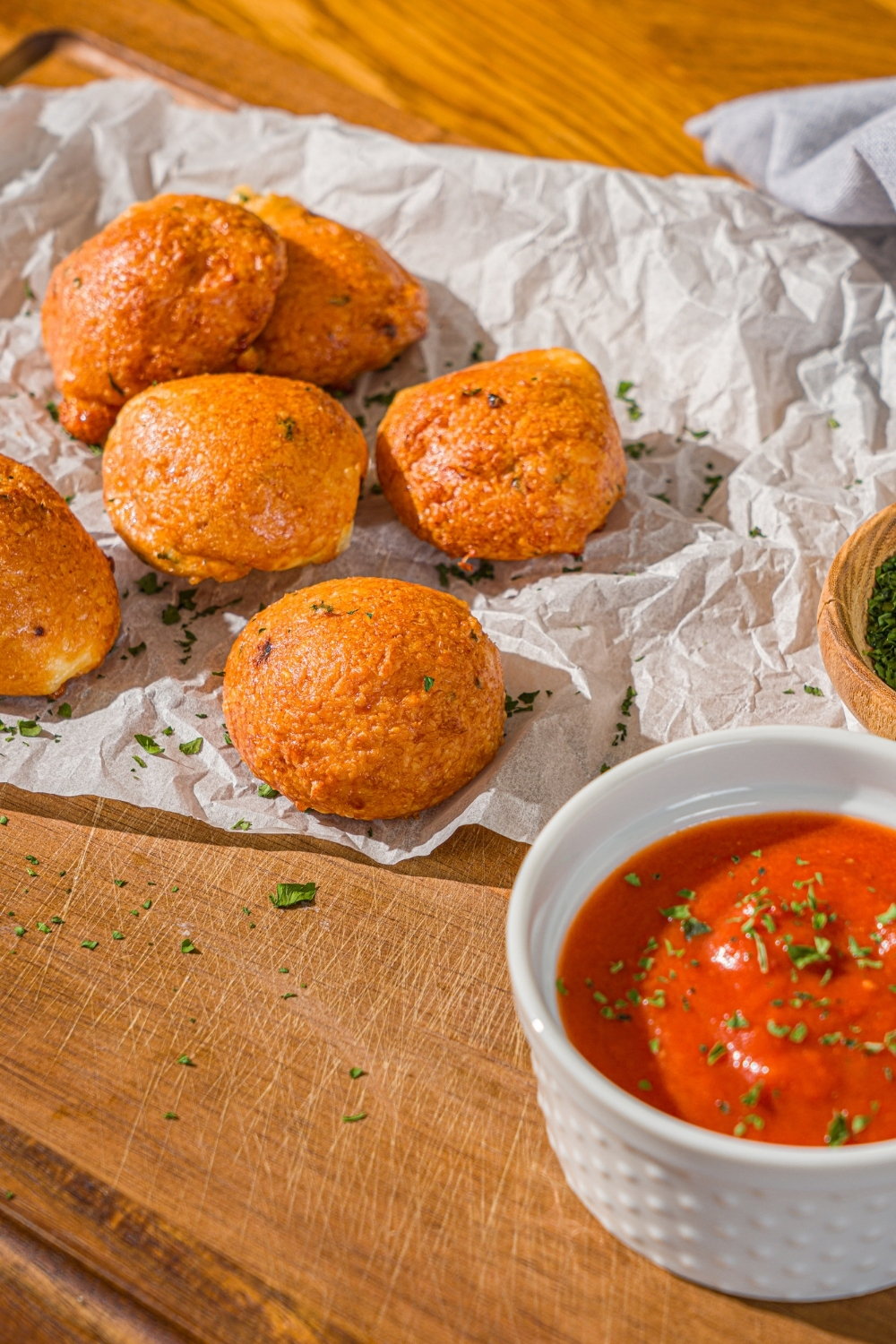 Several air fryer parmesan cheese balls on a piece of parchment paper on a wooden counter. There is a small bowl of parsley and ramekin of marinara sauce on the counter.