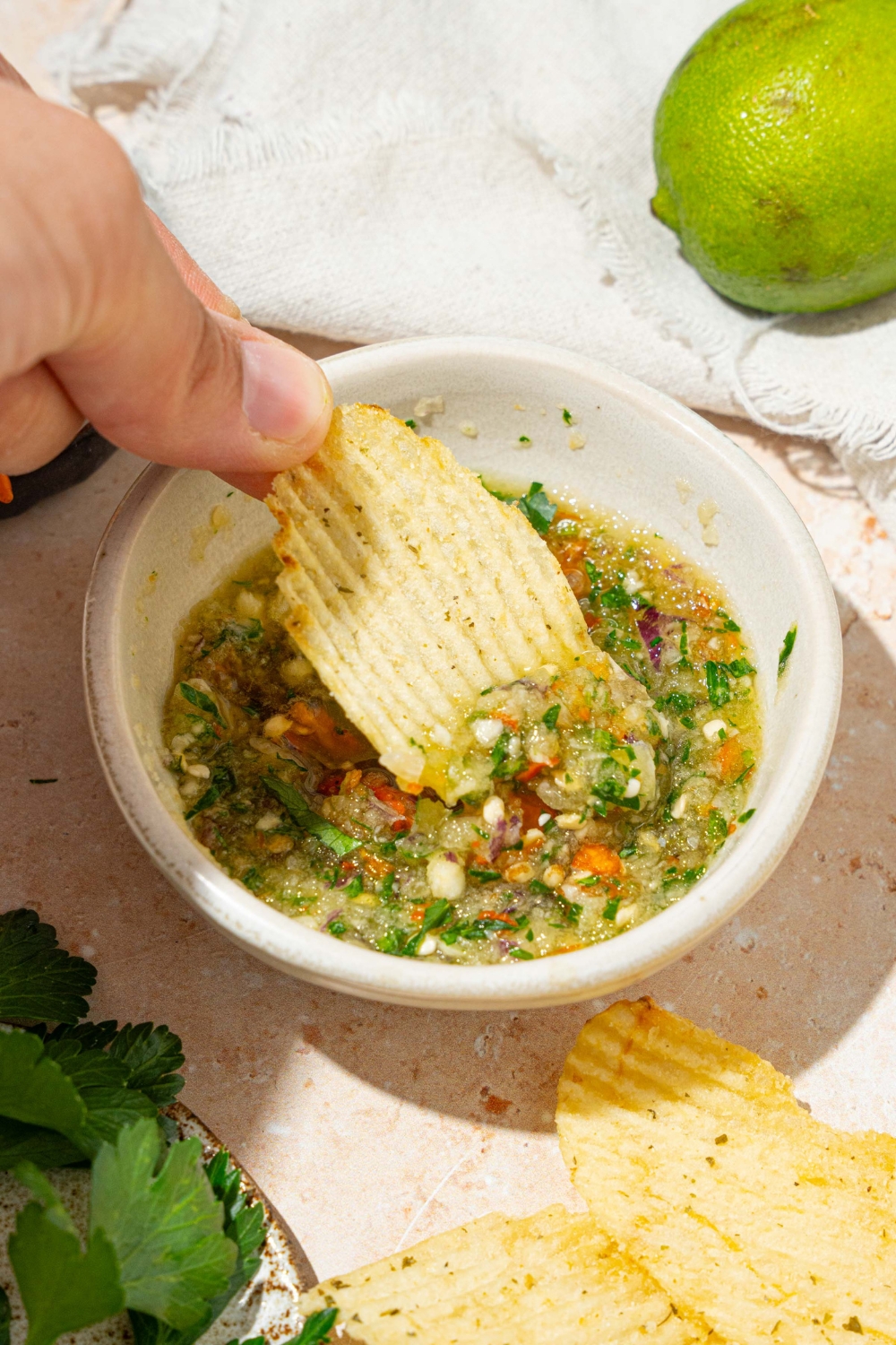 A chip being dipped into a small white bowl with crack sauce. The bowl is on a tan counter with a white cloth napkin and lime.