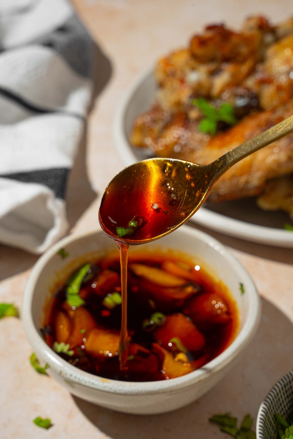 A small bowl of Asian zing sauce with smashed garlic garnished with sliced green onion. There is a spoon dipping in the bowl. The bowl is on a tan counter with a plate of baked wings and small bowls of garnishes.