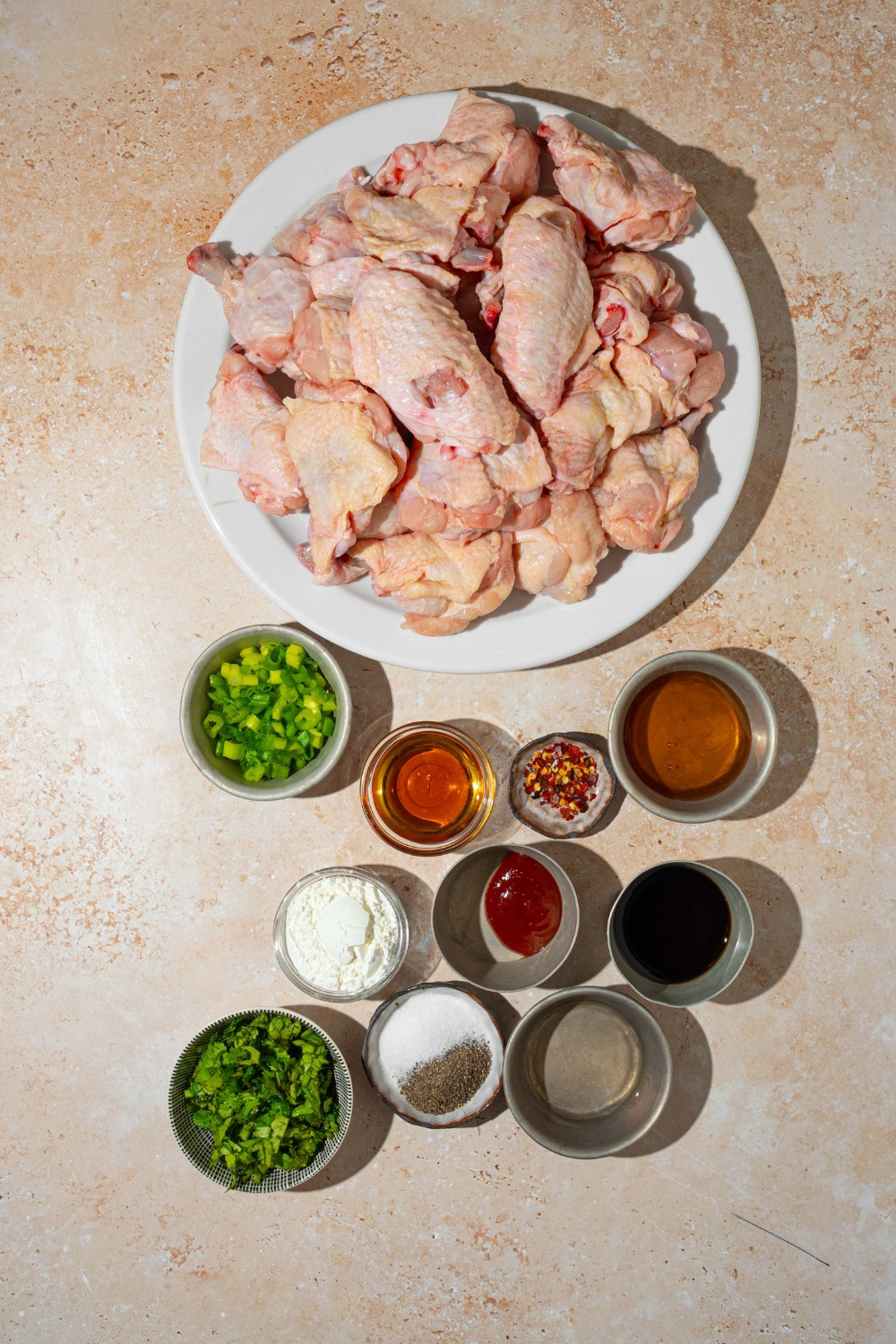A tan counter with several bowls in various sizes containing ingredients to make Asian zing sauce including chicken wings, garlic, soy sauce, honey, mirin, Sriracha, corn starch, chili flakes, sliced green onion, and seasonings.