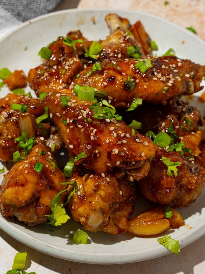 A white plate with baked chicken wings tossed in Asian zing sauce garnished with sesame seeds and sliced green onion. The plate is on a tan counter with a gray cloth napkin.
