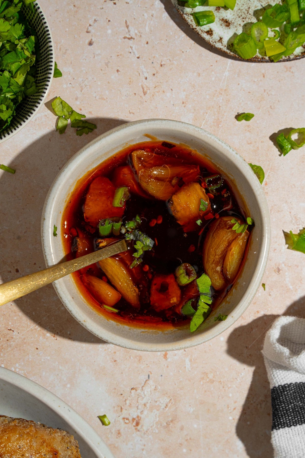 A small white bowl with Asian zing sauce with smashed garlic garnished with sliced green onion. There is a spoon in the bowl. The bowl is on a tan counter with plates of garnishes.