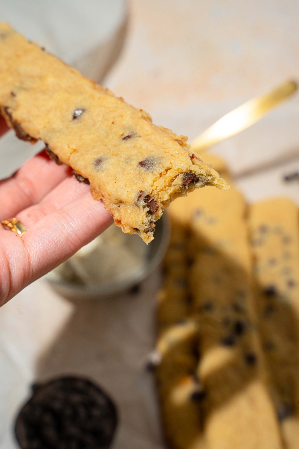 A close up of a chocolate chip cookie fry with a bite taken from the cookie. There is a tan counter with several chocolate chip cookie fries and small bowl of vanilla ice cream blurred in the background.