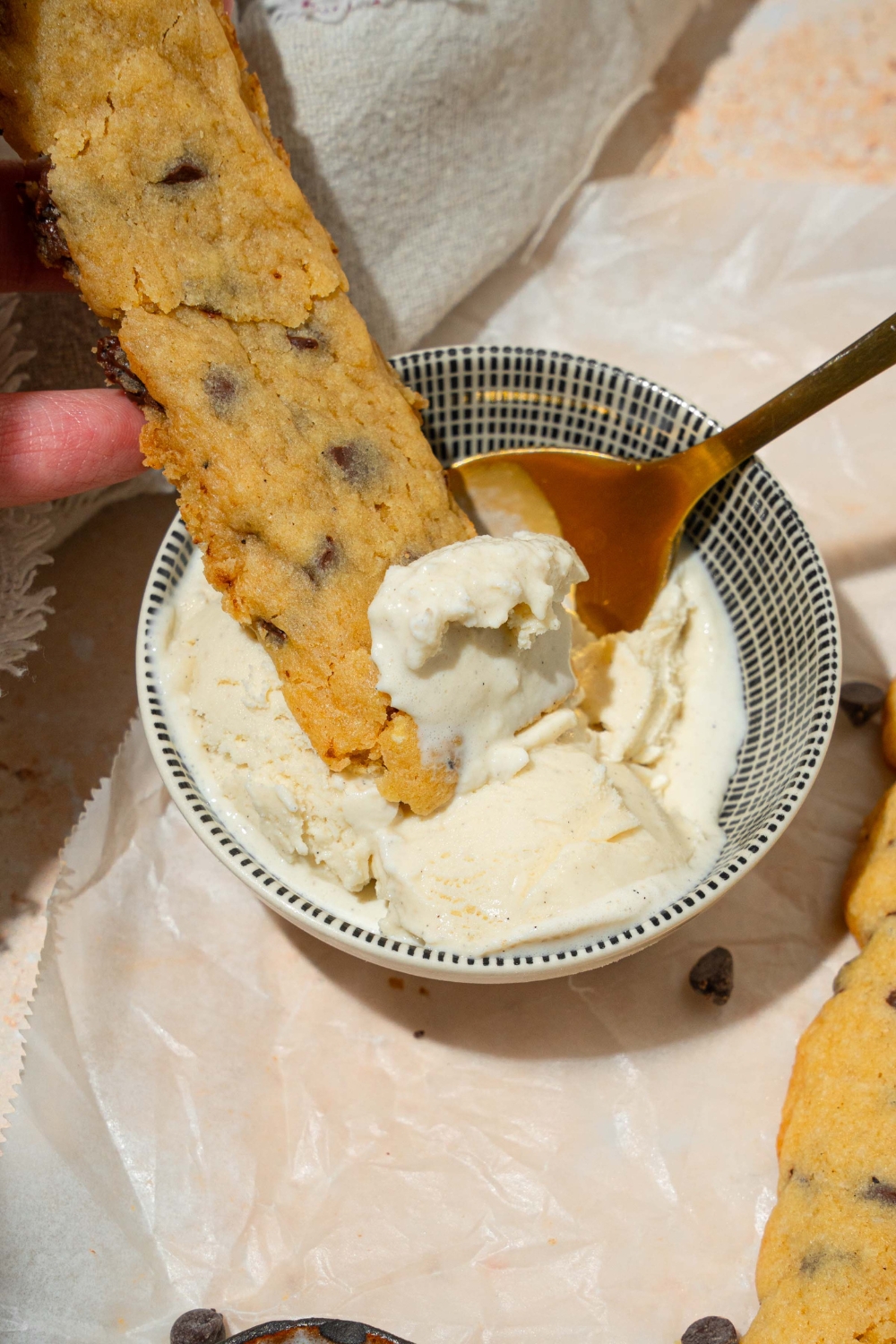 A small bowl of vanilla ice cream with a spoon and a chocolate chip cookie fry dipped in the bowl. The bowl is on a tan counter with chocolate chip cookie fries on a sheet of parchment paper.