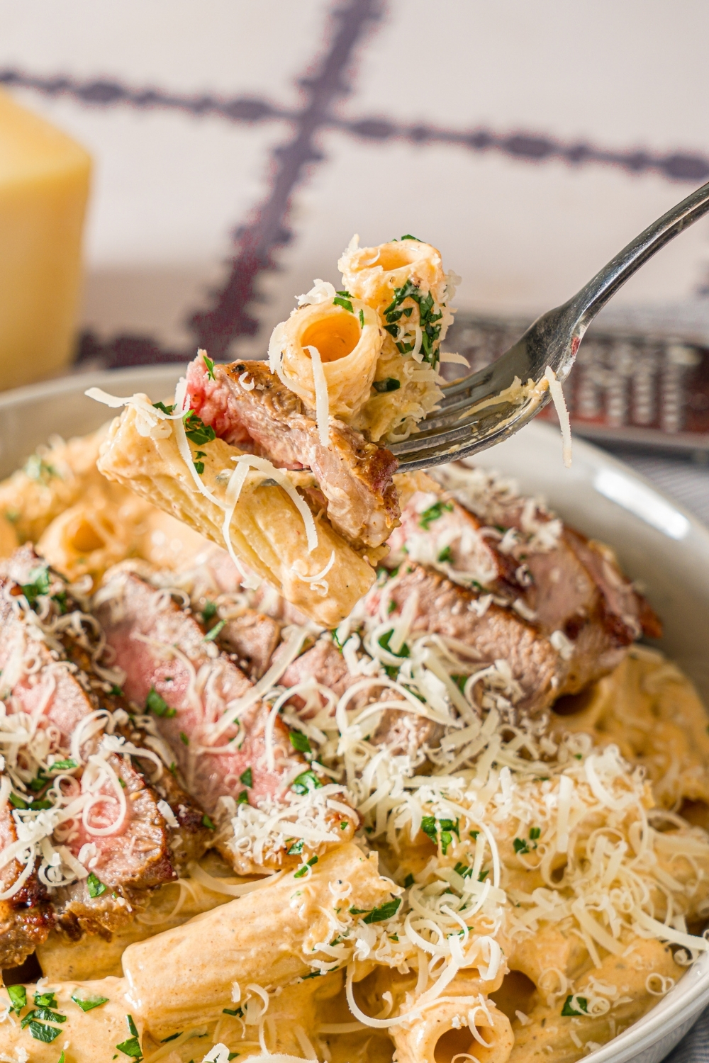 A fork with a bite of creamy cajun steak pasta garnished with fresh parsley and grated cheese. The fork is over a bowl of pasta. The bowl is on a tiled counter with a small bowl of parsley and block of parmesan cheese.