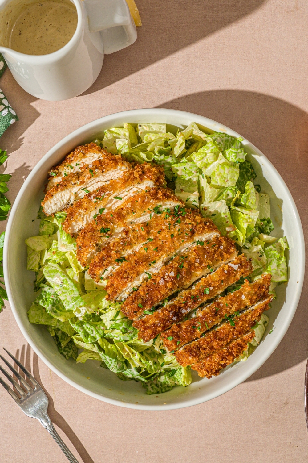 A white bowl with crispy chicken caesar salad garnished with fresh parsley. The bowl is on a tan counter with a fork.