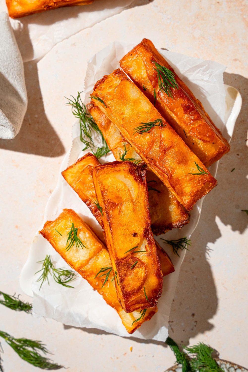 A white platter lined with parchment paper with several pieces of potato pave garnished with fresh thyme. The pieces are stacked on top of each other. The plate is on a tan counter with fresh thyme and a white cloth napkin.