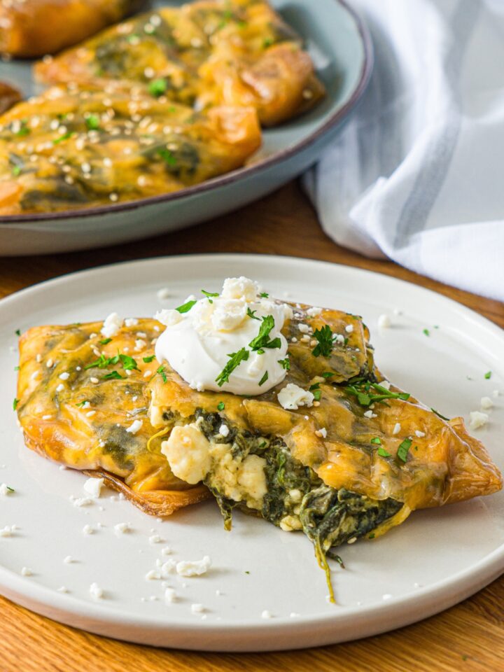 A white plate with rice paper spanakopita split open and topped with sour cream. The plate is garnished with feta cheese and fresh parsley. The plate is on a wooden counter with an additional plate of spanakopita and a white cloth napkin.