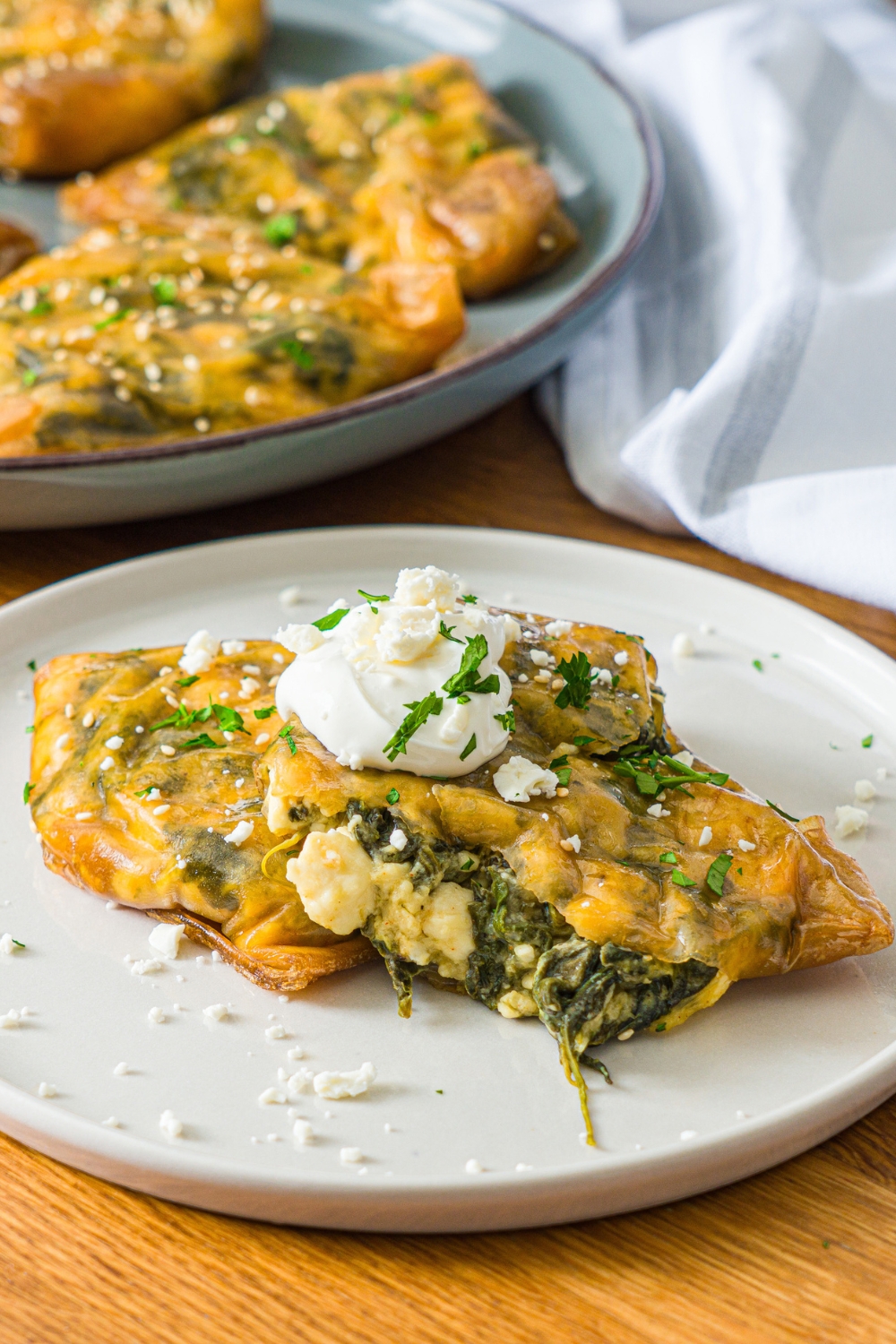 A white plate with rice paper spanakopita split open and topped with sour cream. The plate is garnished with feta cheese and fresh parsley. The plate is on a wooden counter with an additional plate of spanakopita and a white cloth napkin.