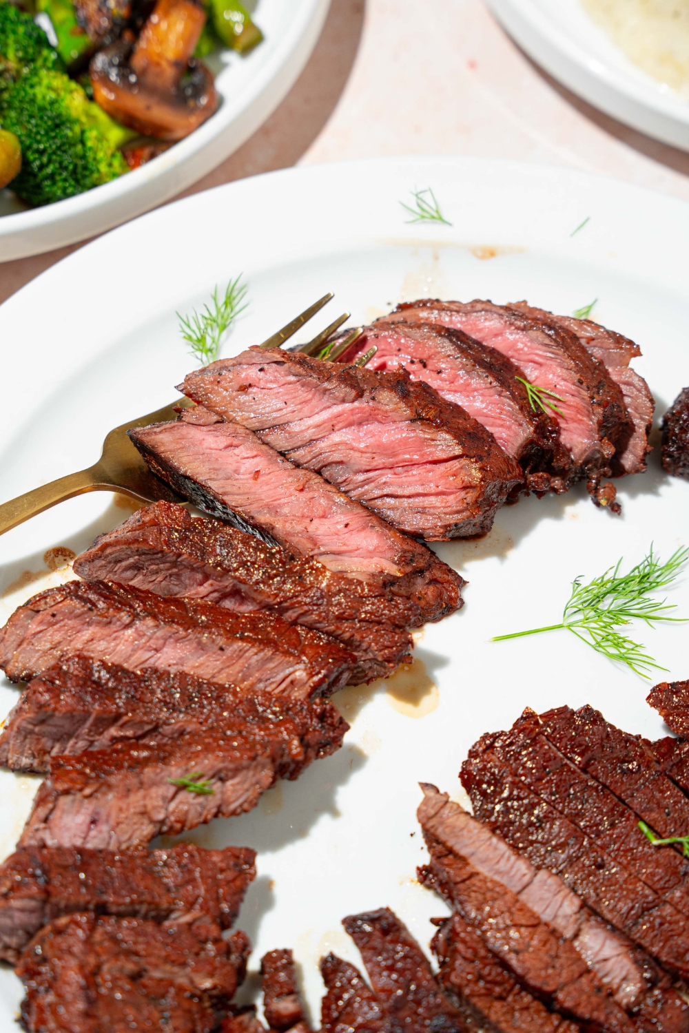A white plate with sliced marinated skirt steak garnished with herbs. There is a fork on the plate. The plate is on a tan counter.