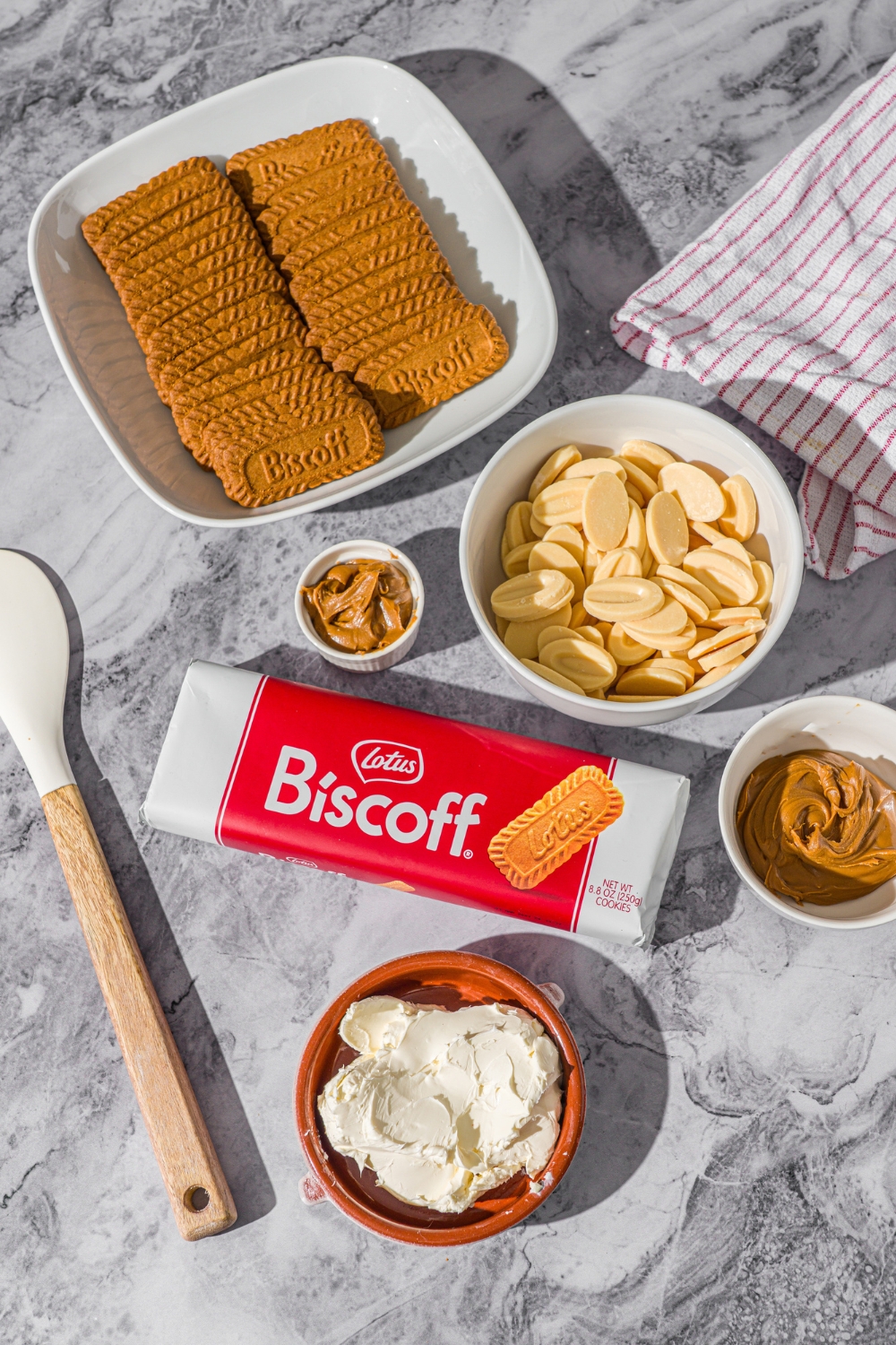 A marble counter with small bowls of various sizes containing ingredients to make biscoff truffles including cream cheese, biscoff cookies, Biscoff cookie butter, and white chocolate.