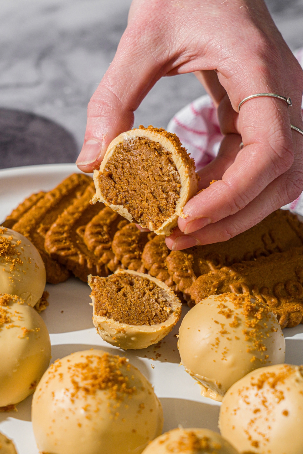 A hand holding a Biscoff truffle sliced in half. The truffles are served with Biscoff cookies. The plate is on a marble counter with a small bowl of cookie crumbs and red striped napkin.