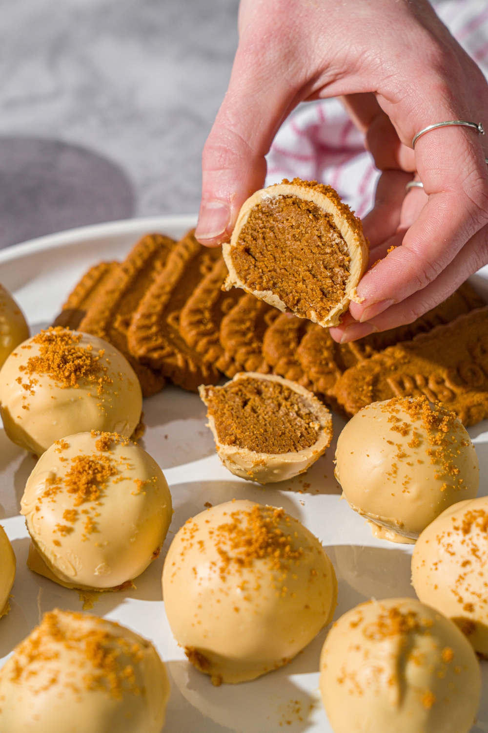 A hand holding a Biscoff truffle sliced in half. The truffle is on a white plate with Biscoff truffles coated in white chocolate and topped with cookie crumbs. The plate is on a marble counter.