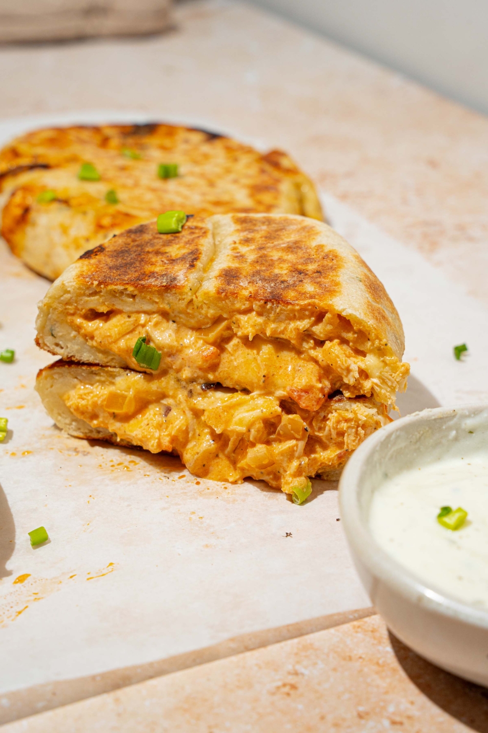 A piece of parchment paper with several Buffalo chicken hot pockets garnished with chives. One of the hot pockets is sliced in half and stacked on one another. The paper is on a tan counter with a bowl of dipping sauce.