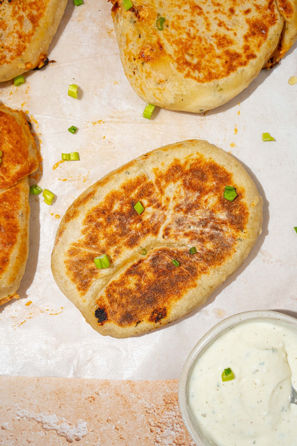 A piece of parchment paper with several Buffalo chicken hot pockets garnished with chives. The paper is on a tan counter with a bowl of dipping sauce.