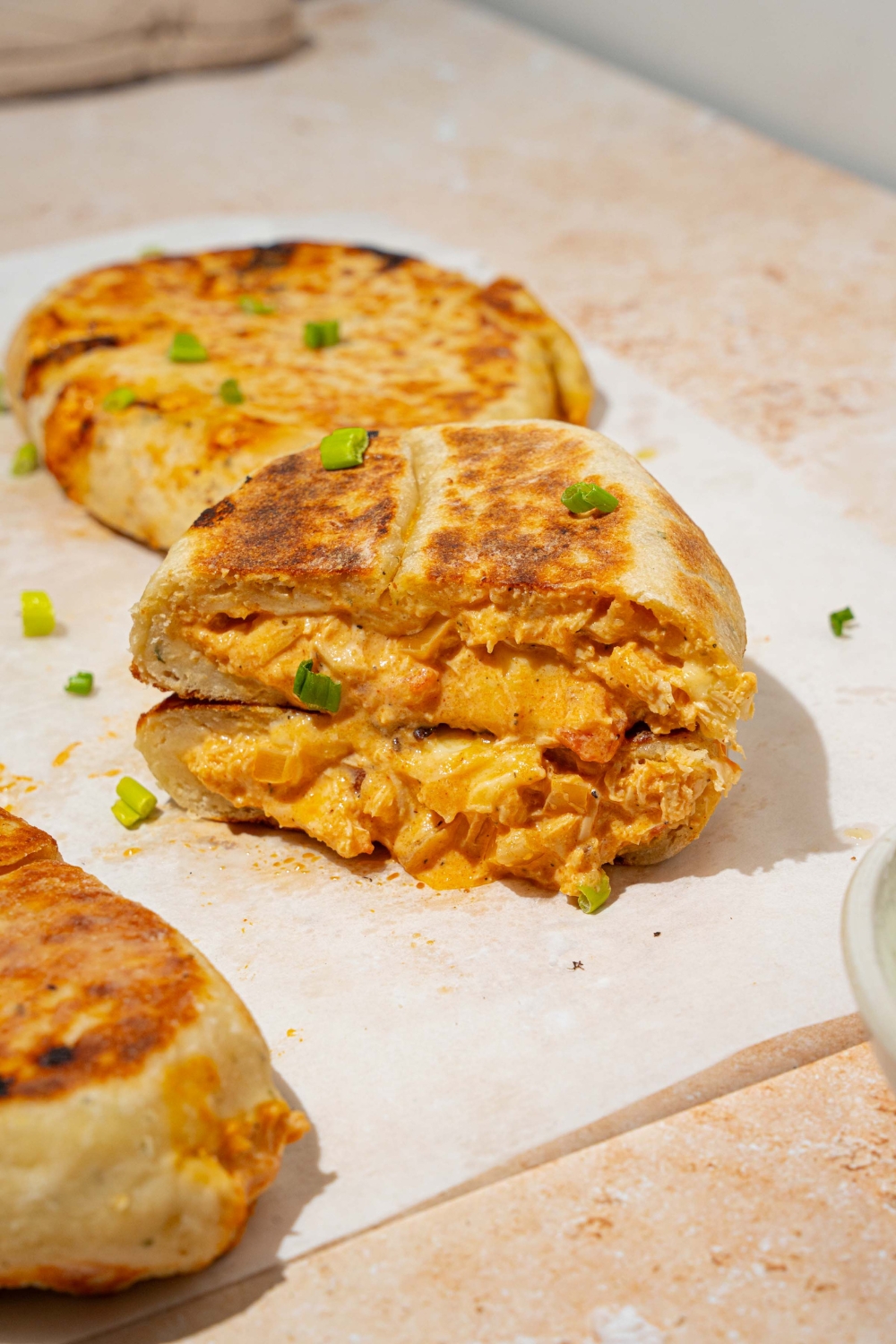 A piece of parchment paper with several Buffalo chicken hot pockets garnished with chives. One of the hot pockets is sliced in half and stacked on one another. The paper is on a tan counter with a bowl of dipping sauce.