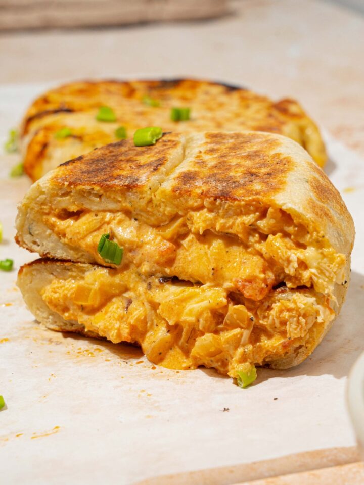A piece of parchment paper with several Buffalo chicken hot pockets garnished with chives. One of the hot pockets is sliced in half and stacked on one another. The paper is on a tan counter with a bowl of dipping sauce.