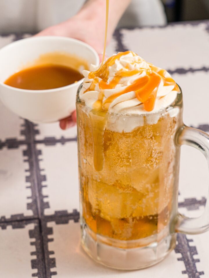 A large beer glass with butterbeer topped with butterscotch whipped cream. Butterscotch syrup is being drizzled over the glass. The glass is on a tiled counter with a small bowl of butterscotch syrup.
