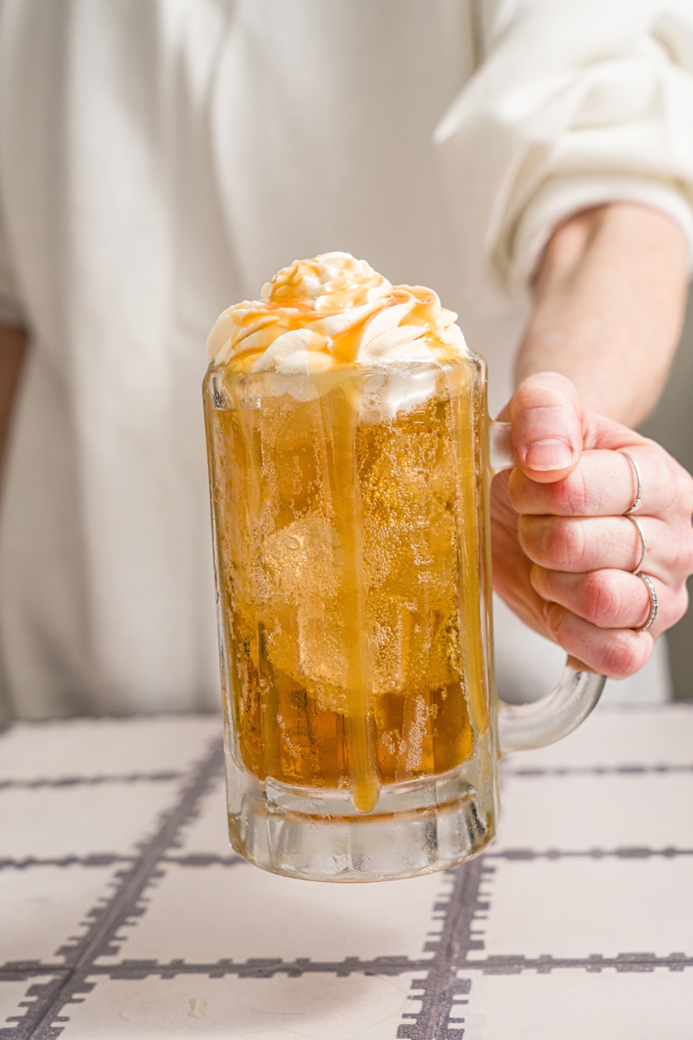 A hand holding a large beer glass with Harry Potter butterbeer topped with butterscotch whipped cream and drizzled with butterscotch syrup.