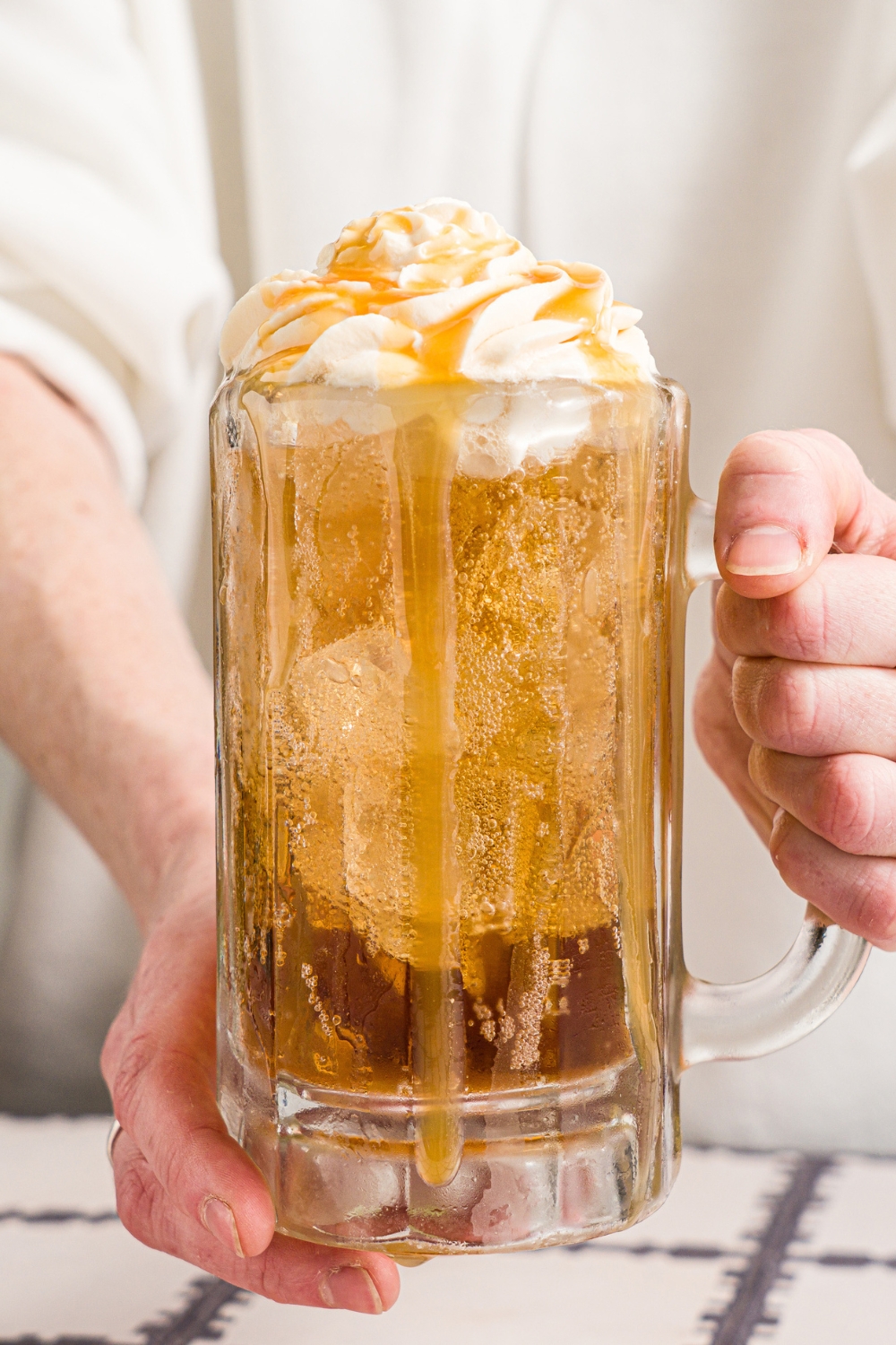 A pair of hands holding a large beer glass with Harry Potter butterbeer topped with butterscotch whipped cream and drizzled with butterscotch syrup.