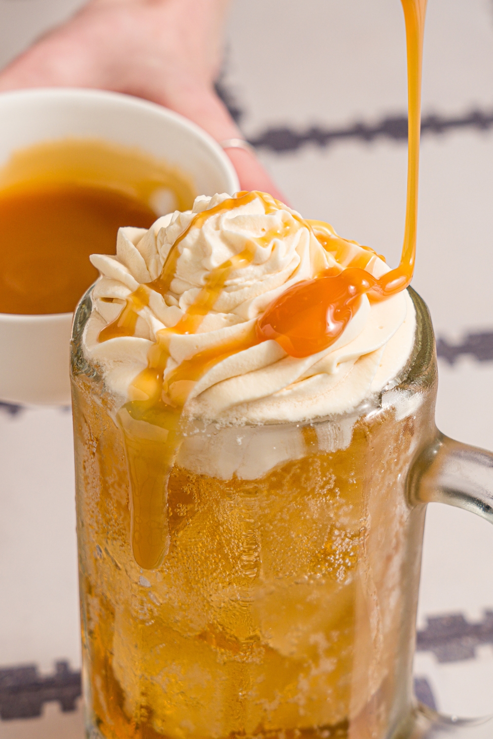 A large beer glass with butterbeer topped with butterscotch whipped cream. Butterscotch syrup is being drizzled over the glass. The glass is on a tiled counter with a small bowl of butterscotch syrup.