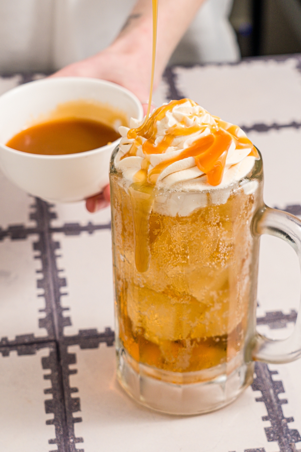 A large beer glass with butterbeer topped with butterscotch whipped cream. Butterscotch syrup is being drizzled over the glass. The glass is on a tiled counter with a small bowl of butterscotch syrup.