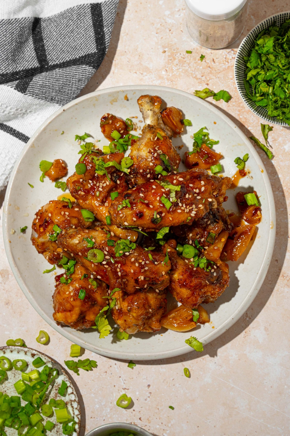 A white plate with baked chicken wings tossed in Asian zing sauce garnished with sesame seeds and sliced green onion. The plate is on a tan counter with a gray cloth napkin.