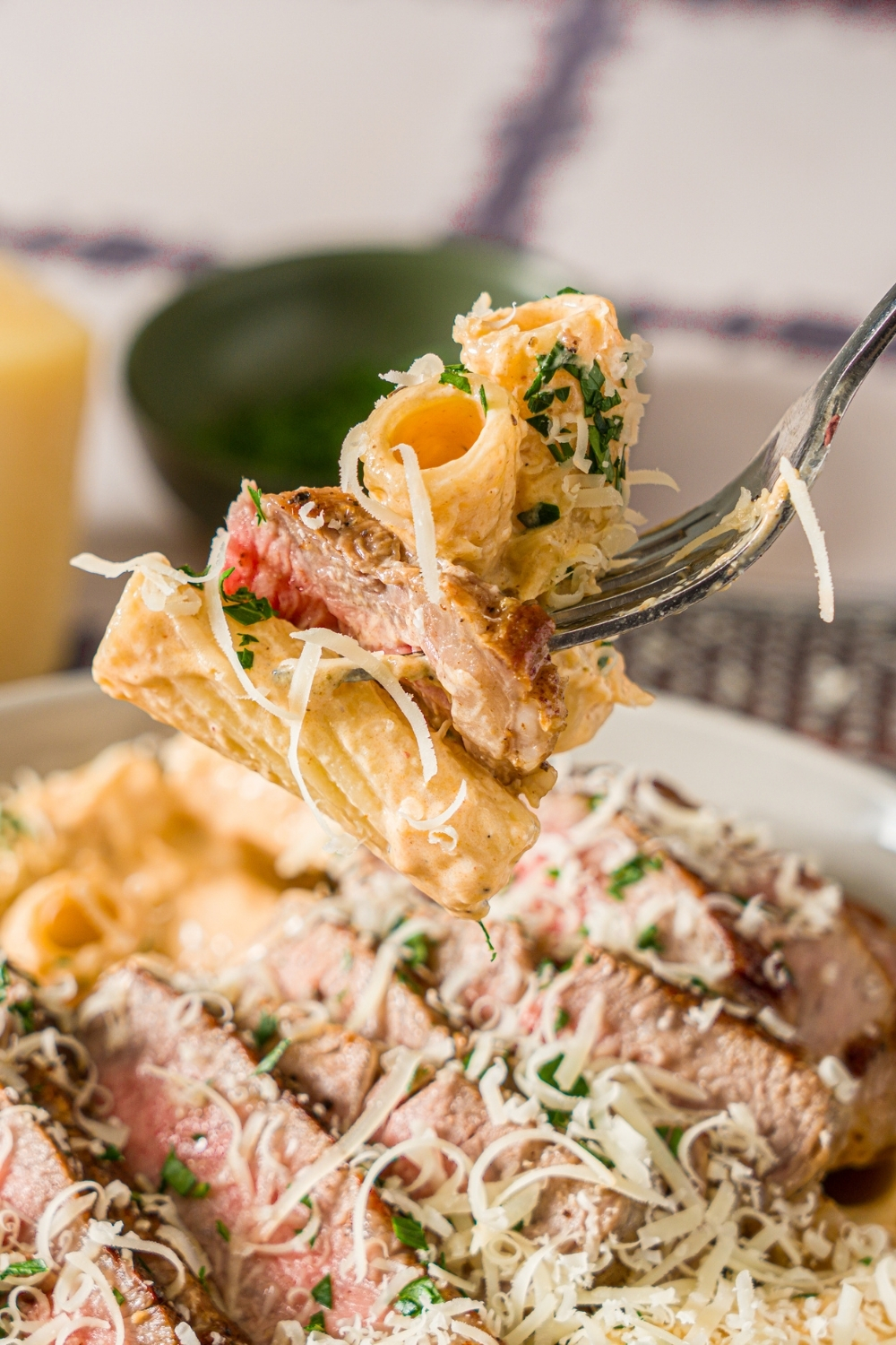 A fork with a bite of creamy cajun steak pasta garnished with fresh parsley and grated cheese. The fork is over a bowl of pasta. The bowl is on a tiled counter with a small bowl of parsley and block of parmesan cheese.