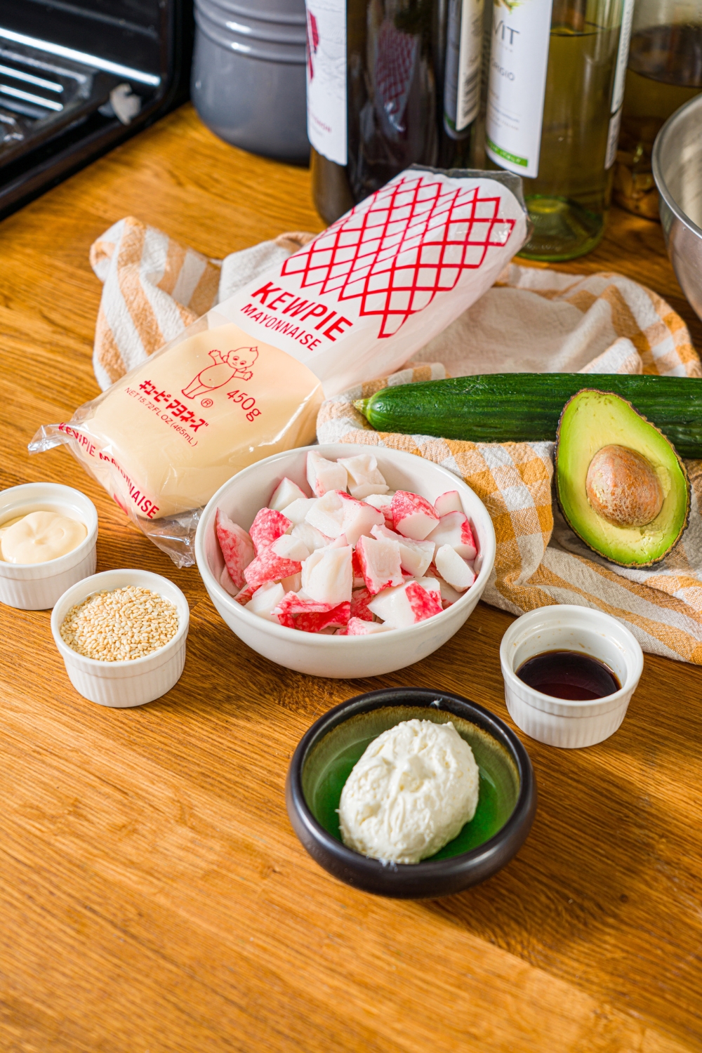 A wooden counter with various bowls containing ingredients to make California roll cucumber salad including cucumber, imitation crab, kewpie mayo, cream cheese, soy sauce, avocado, and sesame seeds.