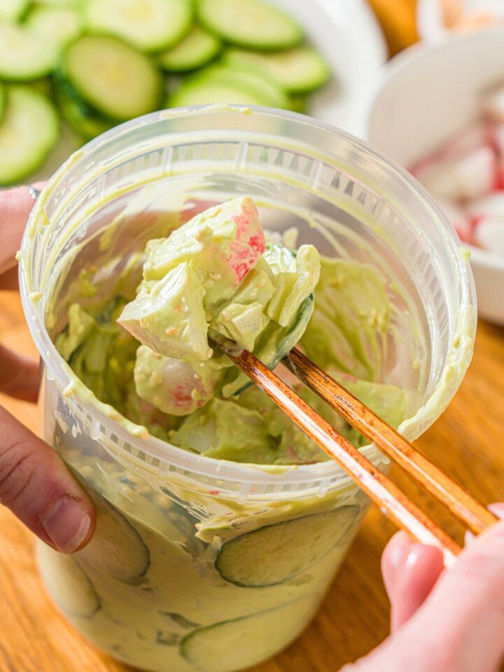 Chopsticks picking up a California roll cucumber salad out of a tub that is filled with the salad