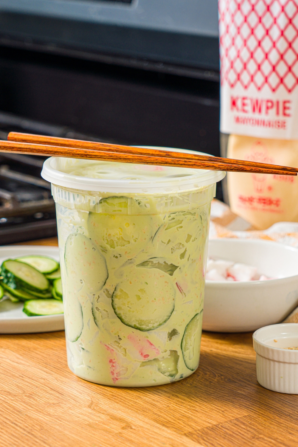 A sealed deli container filled with mixed California roll cucumber salad including sliced cucumber, imitation crab, and sesame seeds in an avocado cream cheese dressing. A pair of chopsticks are on top of the container. The container is on a wooden counter.