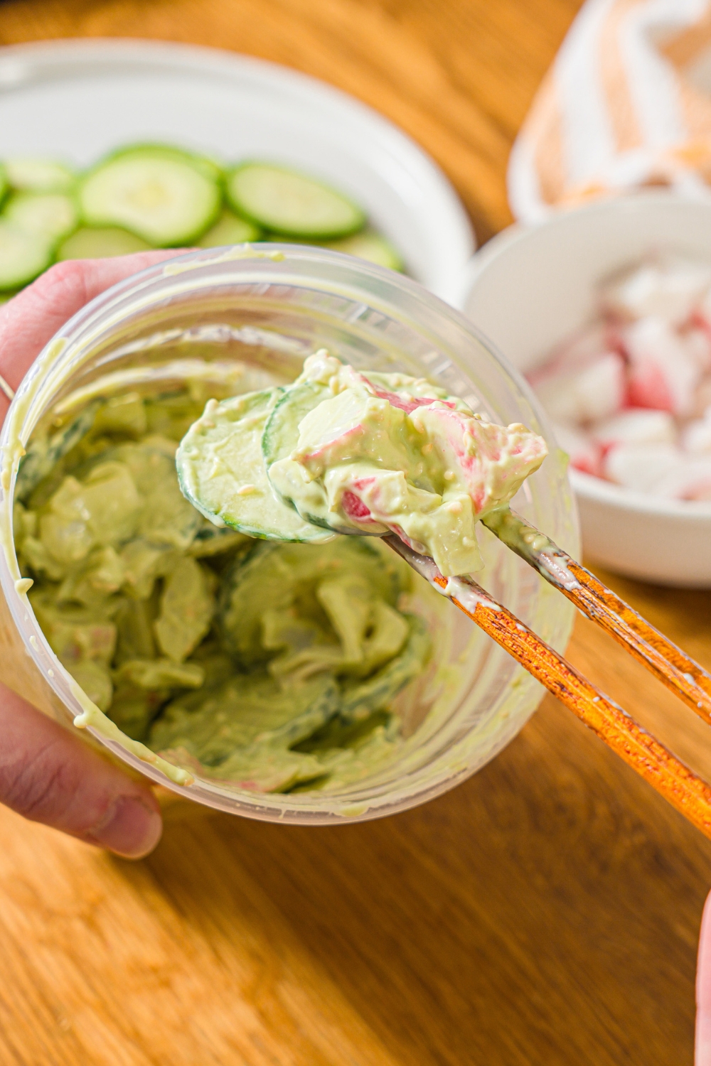 A deli container filled with mixed California roll cucumber salad including sliced cucumber, imitation crab, and sesame seeds in an avocado cream cheese dressing. A pair of chopsticks are taking a bite from the container. The container is on a wooden counter.