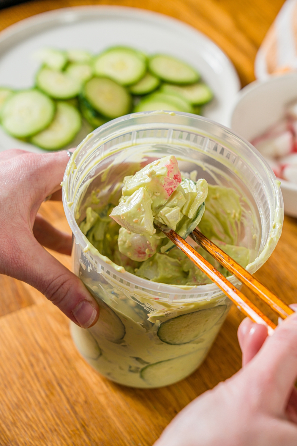 A deli container filled with mixed California roll cucumber salad including sliced cucumber, imitation crab, and sesame seeds in an avocado cream cheese dressing. A pair of chopsticks are taking a bite from the container. The container is on a wooden counter.
