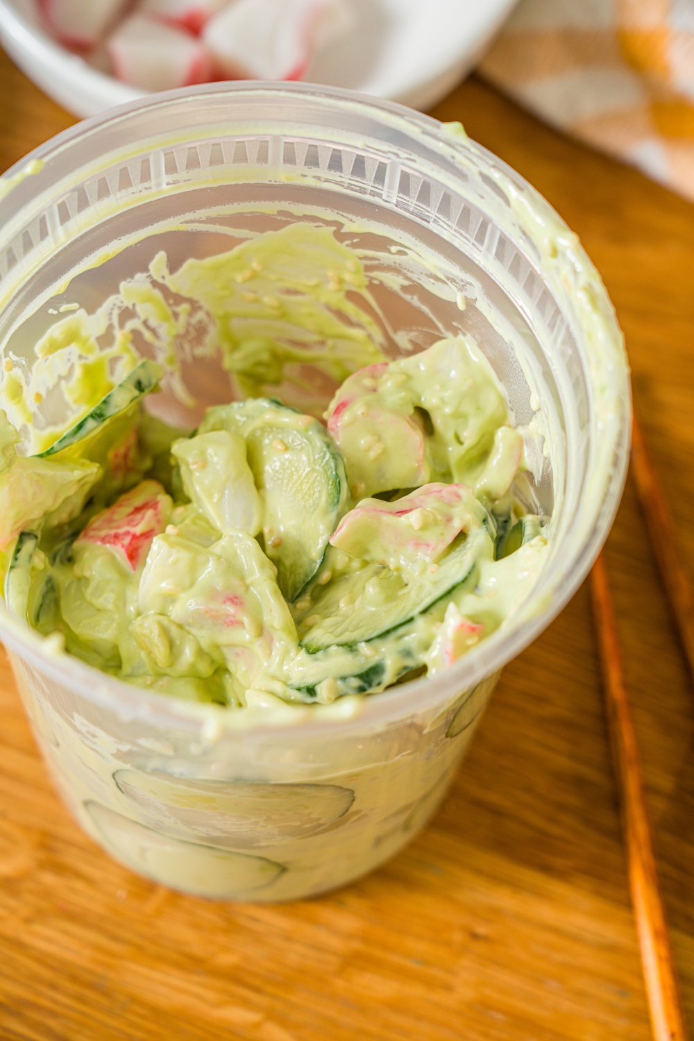 A deli container filled with mixed California roll cucumber salad including sliced cucumber, imitation crab, and sesame seeds in an avocado cream cheese dressing. The container is on a wooden counter with a pair of chopsticks.