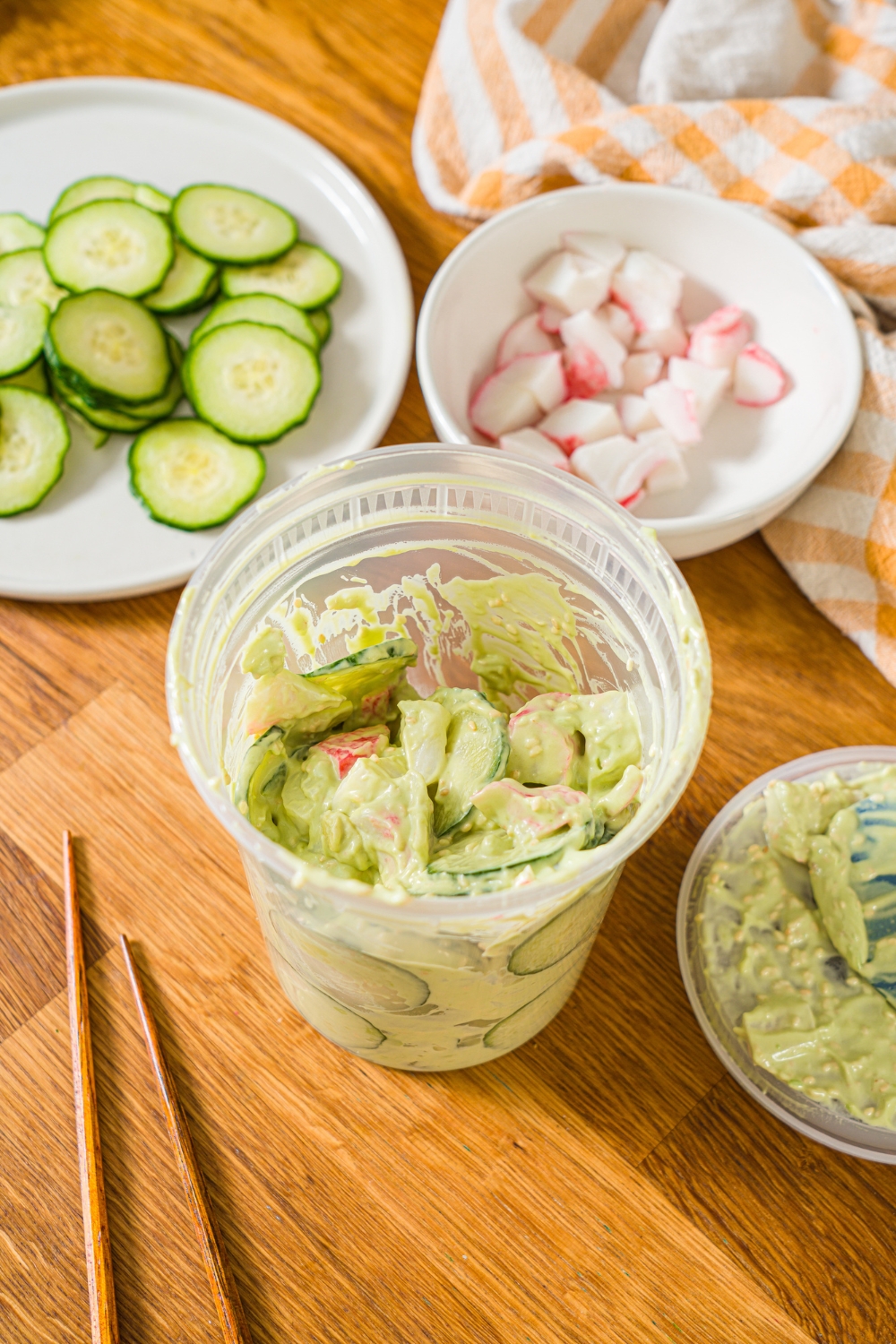A deli container filled with mixed California roll cucumber salad including sliced cucumber, imitation crab, and sesame seeds in an avocado cream cheese dressing. The container is on a wooden counter with a pair of chopsticks and several bowls of ingredients.