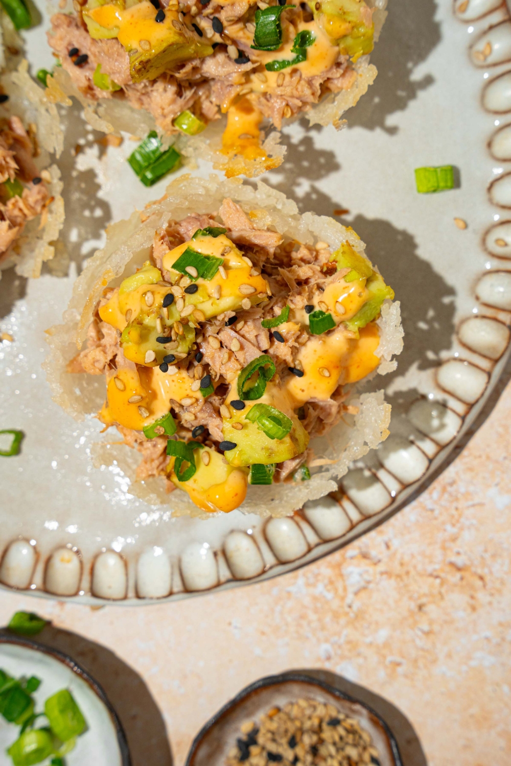 A plate with three tuna rice cups topped with avocado, sliced green onion, sesame seeds, and spicy mayo. The plate is on a tan counter with small plates of garnishes.