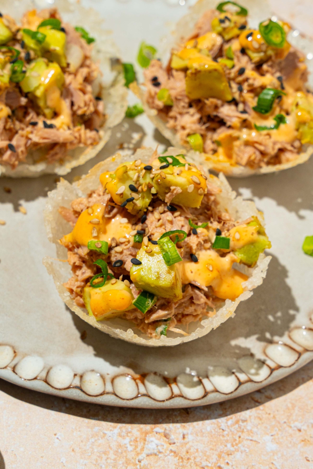 A plate with three tuna rice cups topped with avocado, sliced green onion, sesame seeds, and spicy mayo. The plate is on a tan counter.